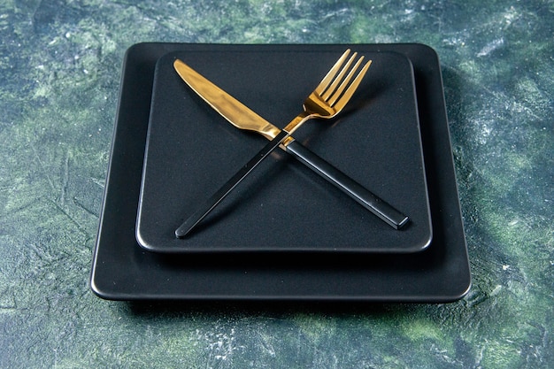 Vista frontal de placas negras con tenedor dorado y cuchillo cruzado sobre fondo oscuro