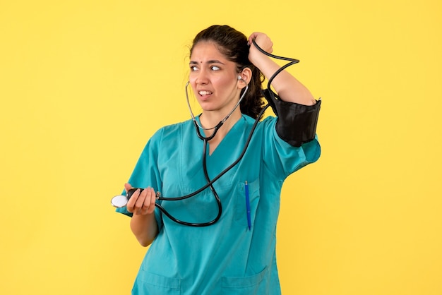 Vista frontal se preguntó doctora en uniforme sosteniendo esfigmomanómetros de pie sobre fondo amarillo