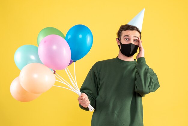 Vista frontal preguntó joven con gorra de fiesta y globos de colores de pie en amarillo