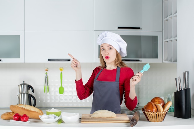 Vista frontal se preguntó mujer rubia con sombrero de cocinero y delantal apuntando a la izquierda en la cocina