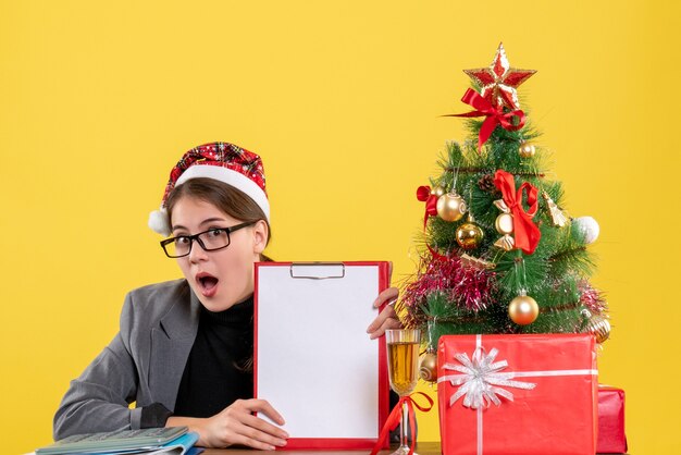 Vista frontal preguntó niña con sombrero de Navidad sentado en la mesa árbol de Navidad y cóctel de regalos