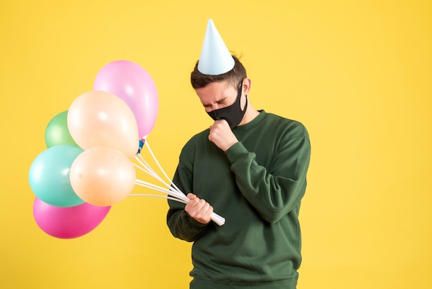 Vista frontal preocupado joven con gorro de fiesta y globos de colores de pie en amarillo