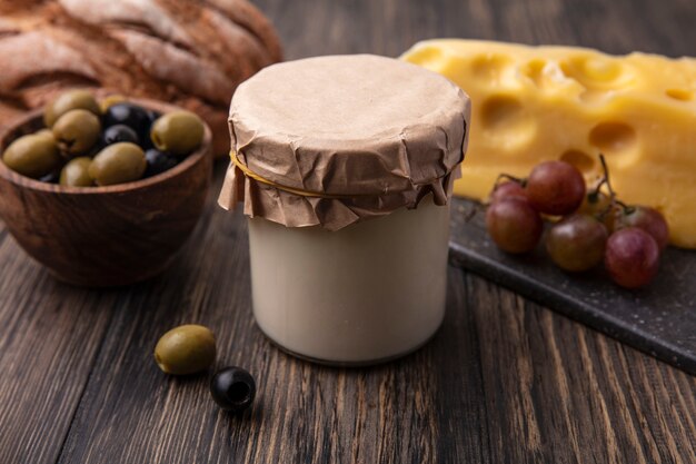 Vista frontal de queso maasdam con uvas en un soporte y aceitunas con yogur en la mesa