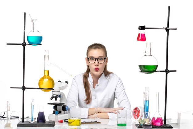 Vista frontal de la química femenina en traje médico frente a la mesa con soluciones sobre fondo blanco química pandémica covid-virus