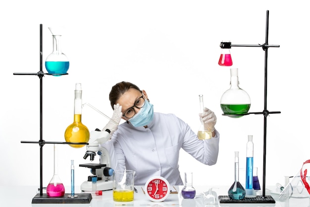 Vista frontal químico femenino en traje médico blanco con máscara que sostiene la solución en el escritorio blanco químico laboratorio virus covid splash
