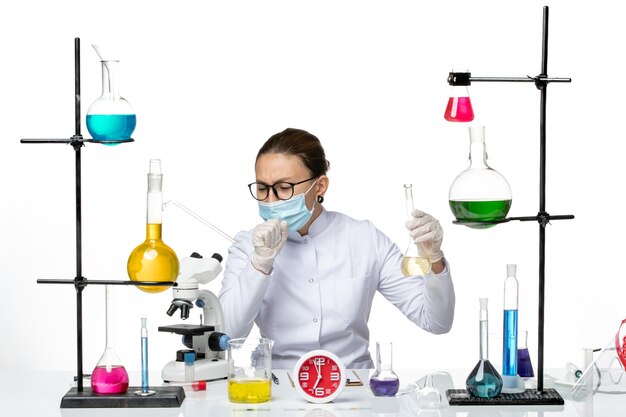 Vista frontal químico femenino en traje médico blanco con máscara que sostiene la solución tosiendo sobre fondo blanco químico laboratorio virus covid- splash