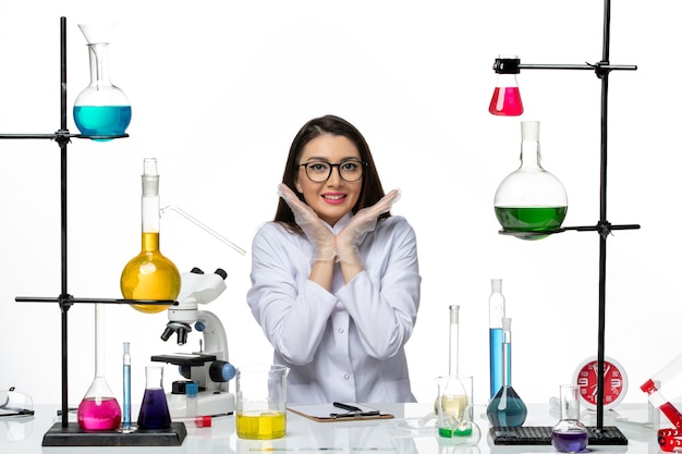 Vista frontal químico femenino en traje médico blanco sentado y posando sonriendo sobre fondo blanco pandemia de covid de virus de ciencia de laboratorio