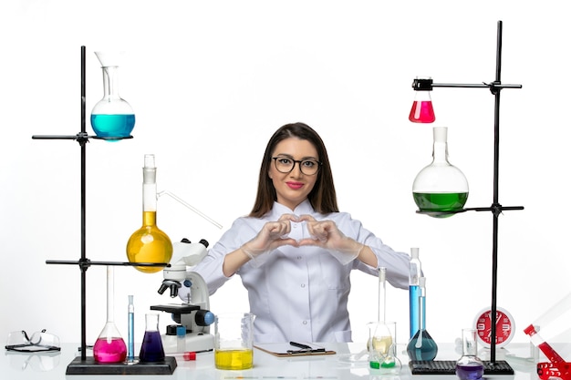 Vista frontal químico femenino en traje médico blanco sentado y sonriendo sobre fondo blanco pandemia covid de virus de ciencia de laboratorio