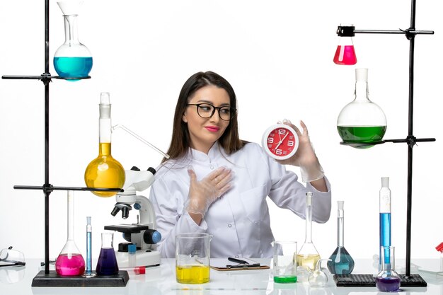 Vista frontal químico femenino en traje médico blanco sosteniendo relojes rojos sobre fondo blanco laboratorio de pandemia de virus de ciencia covid