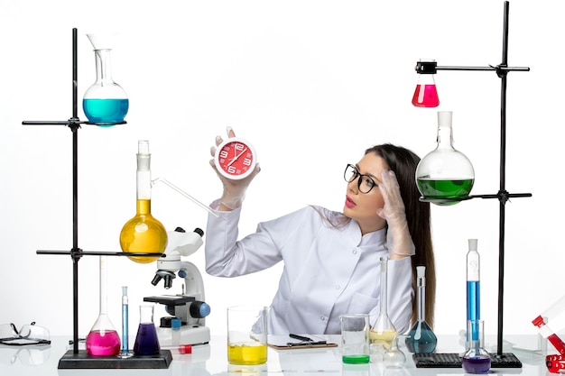 Vista frontal químico femenino en traje médico blanco sosteniendo relojes sobre fondo blanco pandemia de covid de laboratorio de virus de ciencia