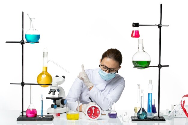 Vista frontal químico femenino en traje médico con máscara sentado con soluciones escribiendo notas sobre fondo blanco químico laboratorio virus covid- splash
