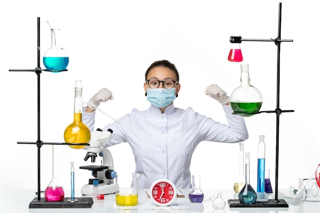 Vista frontal químico femenino en traje médico con máscara sentado con soluciones y flexionando sobre fondo blanco laboratorio de virus química covid splash