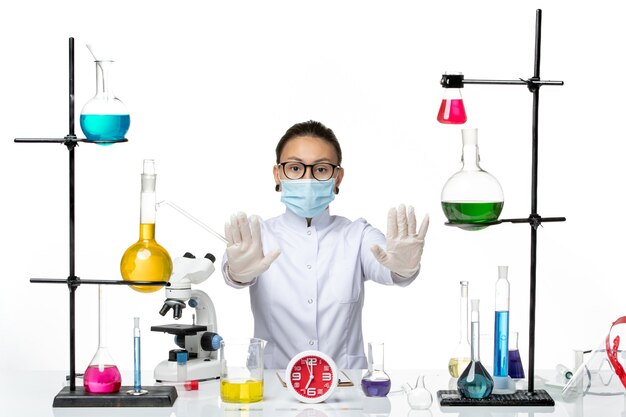 Vista frontal químico femenino en traje médico con máscara sentado con soluciones mirando sobre fondo blanco químico laboratorio virus covid- splash