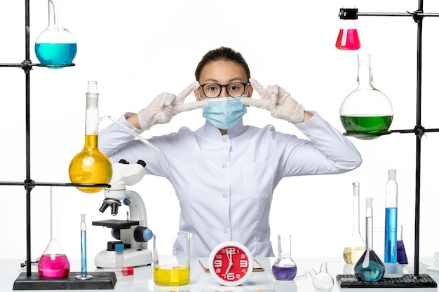 Vista frontal químico femenino en traje médico con máscara sentado con soluciones sobre fondo blanco química virus covid- splash