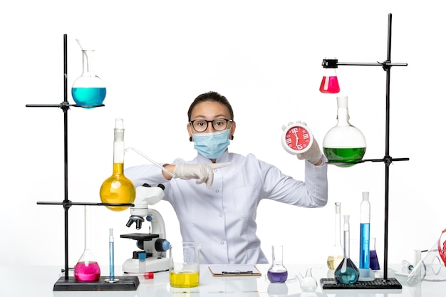 Vista frontal químico femenino en traje médico con máscara sosteniendo relojes en la luz de fondo blanco virus laboratorio química covid splash