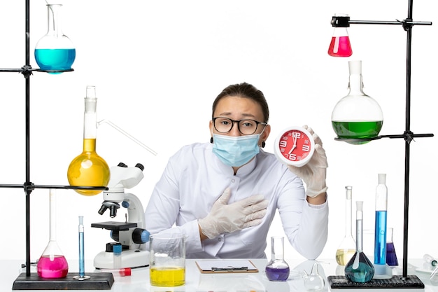 Vista frontal químico femenino en traje médico con máscara sosteniendo relojes rojos sobre fondo blanco química de virus covid- laboratorio de salpicaduras