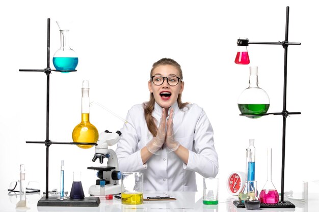 Vista frontal químico femenino en traje médico sentado con soluciones en la química de escritorio blanco covid-virus pandémico