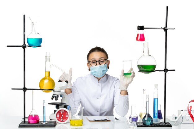 Vista frontal químico femenino en traje médico con solución de sujeción de máscara en el escritorio blanco laboratorio de salpicaduras de química de virus covid-