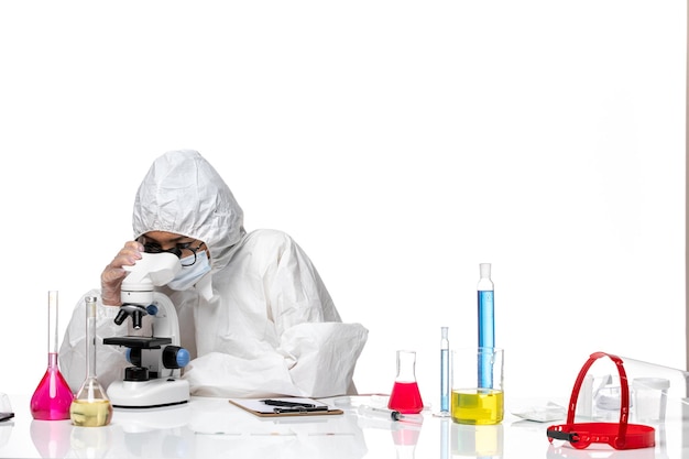 Vista frontal químico femenino en traje de protección especial comprobando la muestra con microscopio sobre fondo blanco química covid-virus de salud pandémica