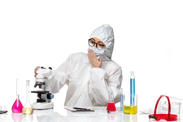 Vista frontal químico femenino en traje de protección especial comprobando la muestra con microscopio sobre fondo blanco química covid- virus de salud