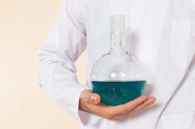 Vista frontal químico masculino en traje especial blanco con matraz con solución azul en el escritorio crema laboratorio de ciencias experimento química