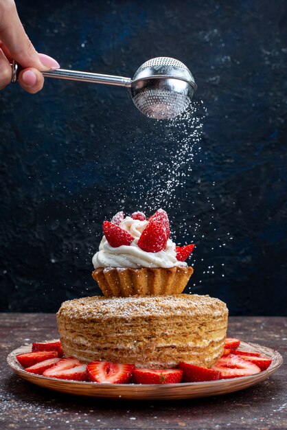Vista frontal de una rebanada de pastel con crema y fresas rojas frescas dentro de la placa obteniendo azúcar en polvo sobre el fondo oscuro