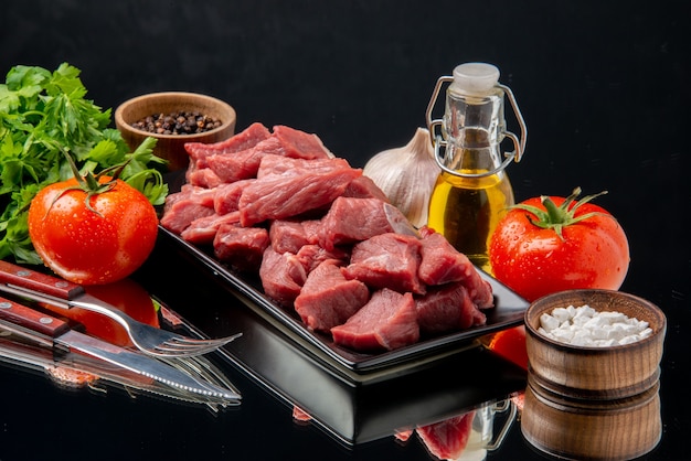 Vista frontal de las rebanadas de carne fresca dentro de la bandeja negra con tomates y verduras sobre una mesa negra