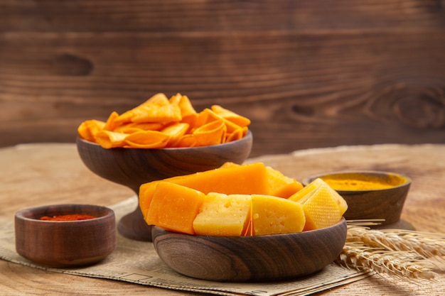 Vista frontal de las rebanadas de queso en un tazón de pimiento rojo en polvo chips espiga de trigo en el periódico en la superficie aislada