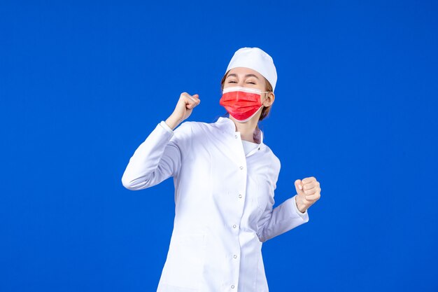 Vista frontal regocijándose joven enfermera en traje médico con máscara protectora roja sobre azul