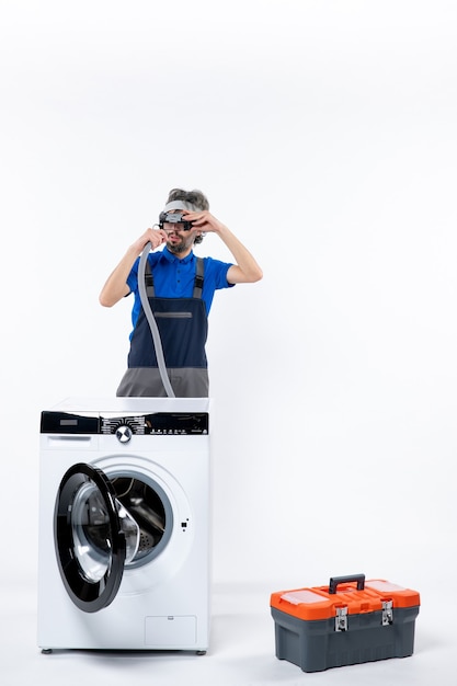 Foto gratuita vista frontal del reparador en uniforme de pie detrás de la lavadora con lámpara de cabeza comprobando el tubo en la pared blanca
