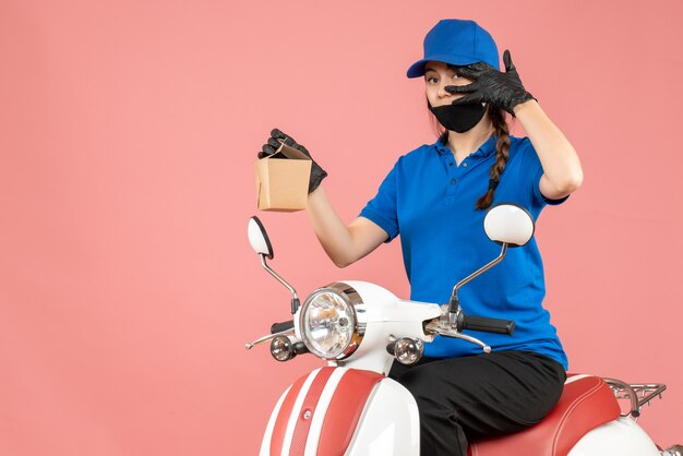 Vista frontal del repartidor confiado con máscara médica y guantes sentado en scooter entregando pedidos sobre fondo melocotón pastel