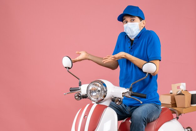 Vista frontal del repartidor confundido con máscara médica con sombrero sentado en scooter sobre fondo de melocotón pastel