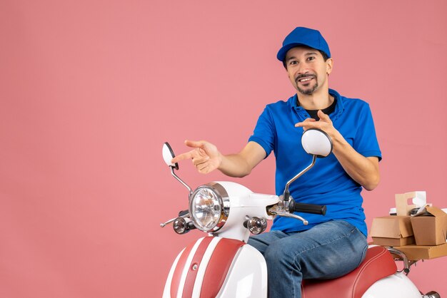 Vista frontal del repartidor feliz con sombrero sentado en scooter sobre fondo de melocotón pastel