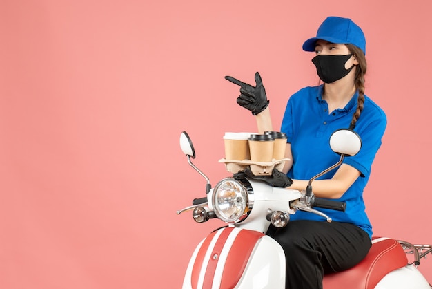 Foto gratuita vista frontal del repartidor femenino confundido con máscara médica y guantes sentado en scooter con órdenes sobre fondo melocotón pastel