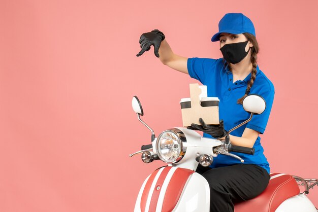 Vista frontal del repartidor femenino emocional con máscara médica y guantes sentado en scooter entregando pedidos sobre fondo melocotón pastel