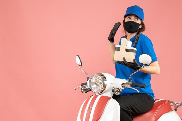 Vista frontal del repartidor femenino enfocado con máscara médica y guantes sentado en scooter entregando pedidos sobre fondo melocotón pastel