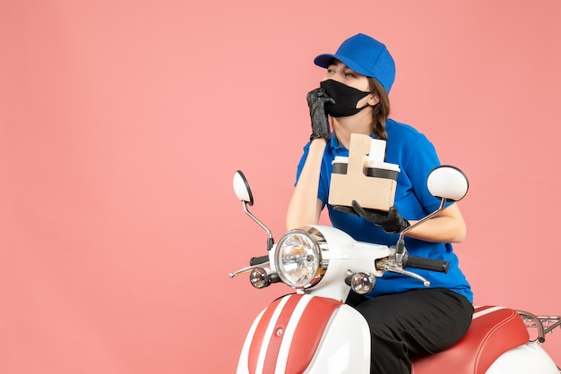 Vista frontal del repartidor femenino pensativo con máscara médica y guantes sentado en scooter entregando pedidos sobre fondo melocotón pastel