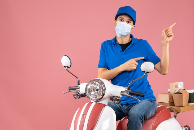 Vista frontal del repartidor con máscara médica con sombrero sentado en scooter apuntando algo en el lado izquierdo sobre fondo melocotón pastel