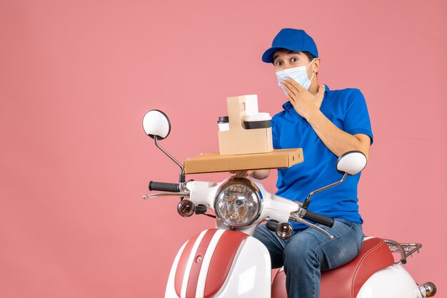 Vista frontal del repartidor masculino interesado en máscara con sombrero sentado en scooter entregando pedidos sobre fondo melocotón pastel