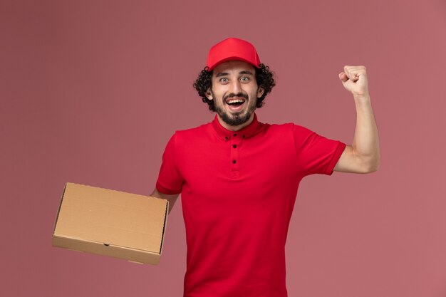Vista frontal repartidor de mensajería masculina en camisa roja y capa con caja de comida de entrega en la pared rosada trabajo masculino empleado de la empresa de entrega de servicios