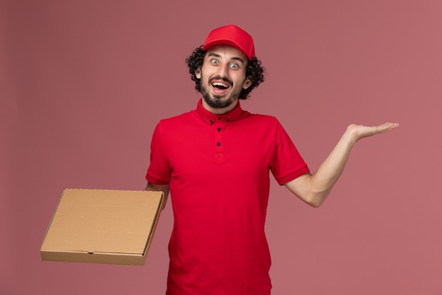 Vista frontal repartidor de mensajería masculino en camisa roja y capa con caja de comida de entrega en la pared rosa empleado de la empresa de entrega de servicios