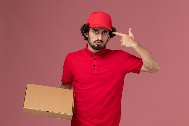 Vista frontal repartidor de mensajería masculino en camisa roja y capa con caja de comida de entrega en la pared rosa trabajador empleado de la empresa de entrega de servicios