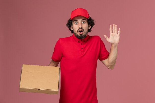 Vista frontal repartidor de mensajería masculino en camisa roja y capa con caja de comida de entrega en el trabajo de empleado de la empresa de entrega de servicio de pared rosa