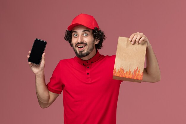 Vista frontal repartidor de mensajería masculino en camisa roja y capa con paquete de alimentos y teléfono inteligente en el empleado de entrega de servicio de pared rosa