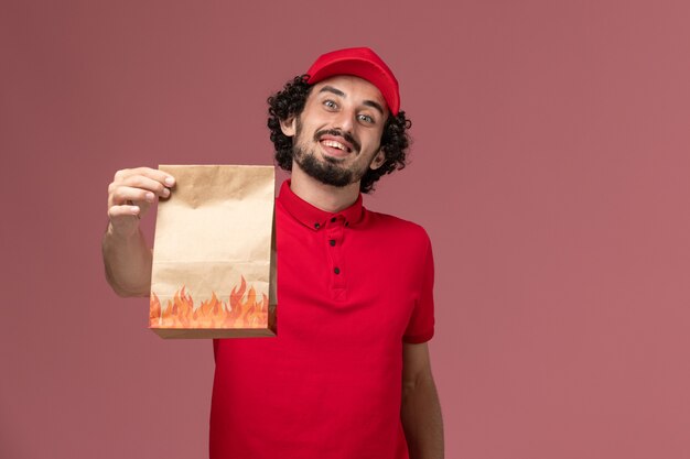 Vista frontal repartidor de mensajería masculino en camisa roja y capa con paquete de comida de papel en la pared rosa empleado de la empresa de entrega de servicios de trabajo