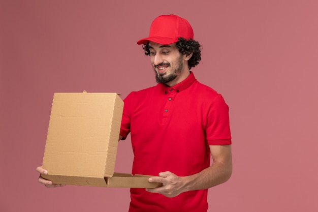 Vista frontal repartidor de mensajería masculino con camisa roja y capa sosteniendo una caja de comida vacía en el trabajo de empleado de la empresa de entrega de servicios de pared rosa