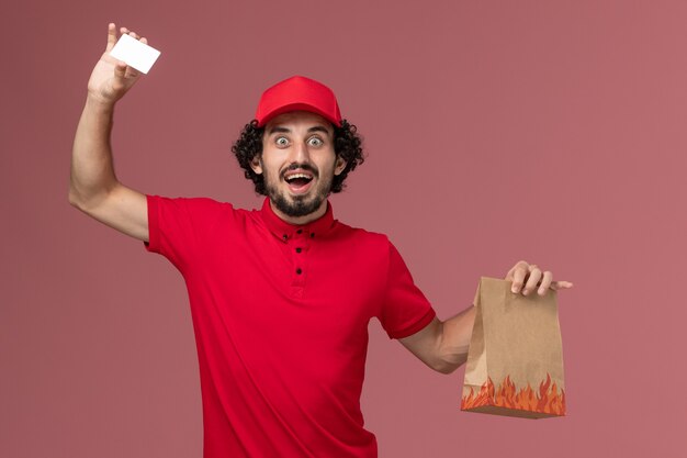 Vista frontal repartidor de mensajería masculino en camisa roja y capa sosteniendo el paquete de alimentos y la tarjeta en el trabajo de empleado de entrega de servicio de pared rosa