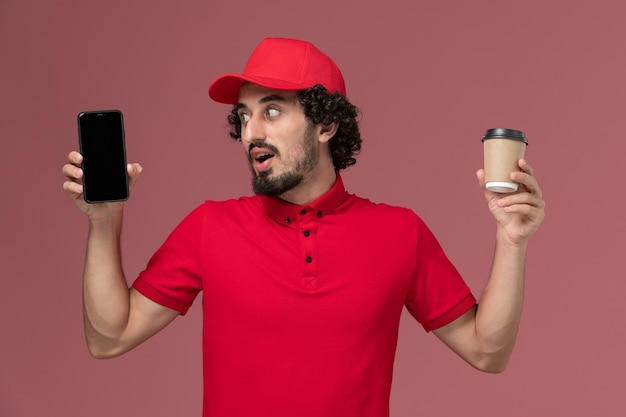 Vista frontal repartidor de mensajería masculino en camisa roja y capa sosteniendo una taza de café de entrega marrón y un teléfono en el trabajo de empleado de servicio de entrega de pared rosa claro