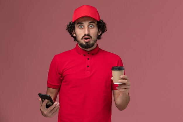 Vista frontal repartidor de mensajería masculino en camisa roja y capa sosteniendo una taza de café de entrega marrón y usando su teléfono en la pared rosa claro empleado de servicio de entrega