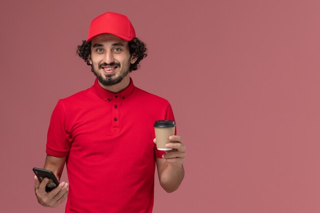 Vista frontal repartidor de mensajería masculino con camisa roja y capa sosteniendo una taza de café de entrega marrón y usando el teléfono en la pared rosa claro empleado de servicio de entrega
